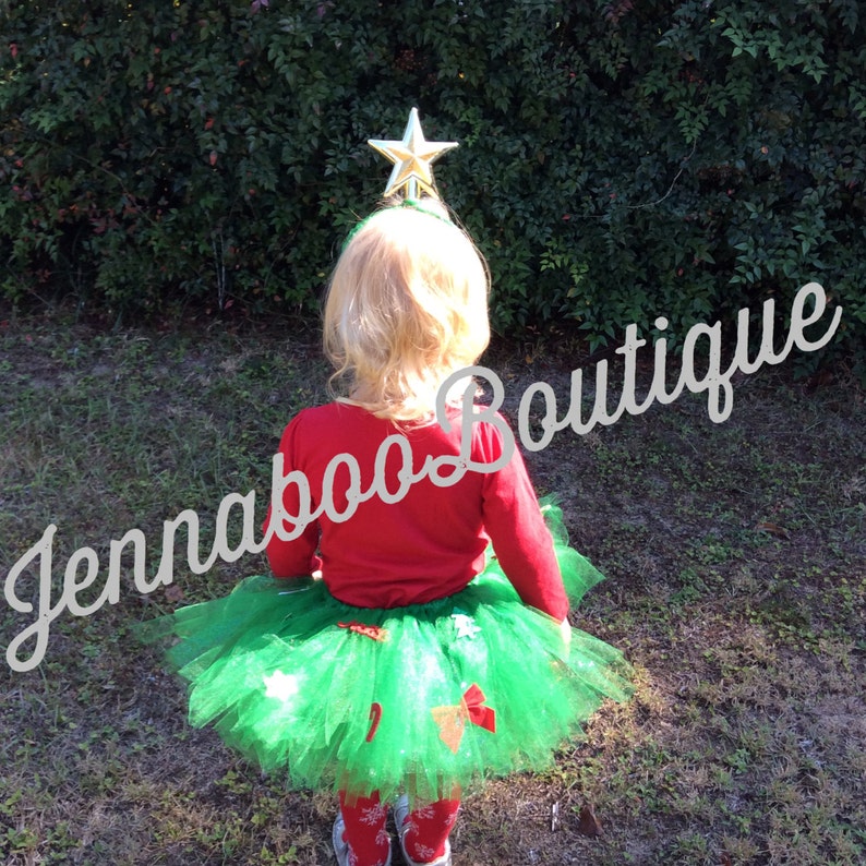 May include: A child wearing a green tulle skirt with red and green Christmas tree decorations. The skirt is paired with a red long-sleeved shirt and a gold star headband. The child is standing in front of a green bush.