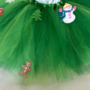 May include: Green tulle tutu skirt with festive felt appliques including a snowman, gingerbread people, a Christmas tree, and a snowflake.