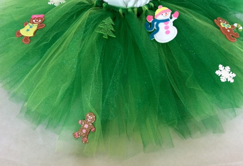 May include: Green tulle tutu skirt with festive felt appliques including a snowman, gingerbread people, a Christmas tree, and a snowflake.