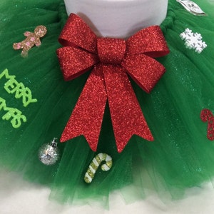 May include: A green tulle tutu with a red glitter bow and various Christmas-themed decorations, including a gingerbread man, a snowflake, a candy cane, and an ornament. The tutu has the words "Merry Christmas" written on it.