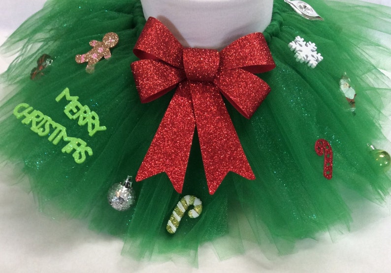 May include: A green tulle tutu with a red glitter bow and various Christmas-themed decorations, including a gingerbread man, a snowflake, a candy cane, and an ornament. The tutu has the words "Merry Christmas" written on it.