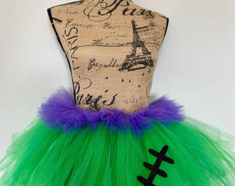 May include: Green and purple tulle tutu with black glitter stitches on the front. The tutu is on a dress form.