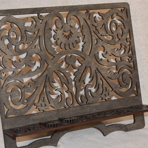May include: A dark brown wooden book stand with a decorative, intricate, laser-cut design. The stand has a shelf for holding books or other items.