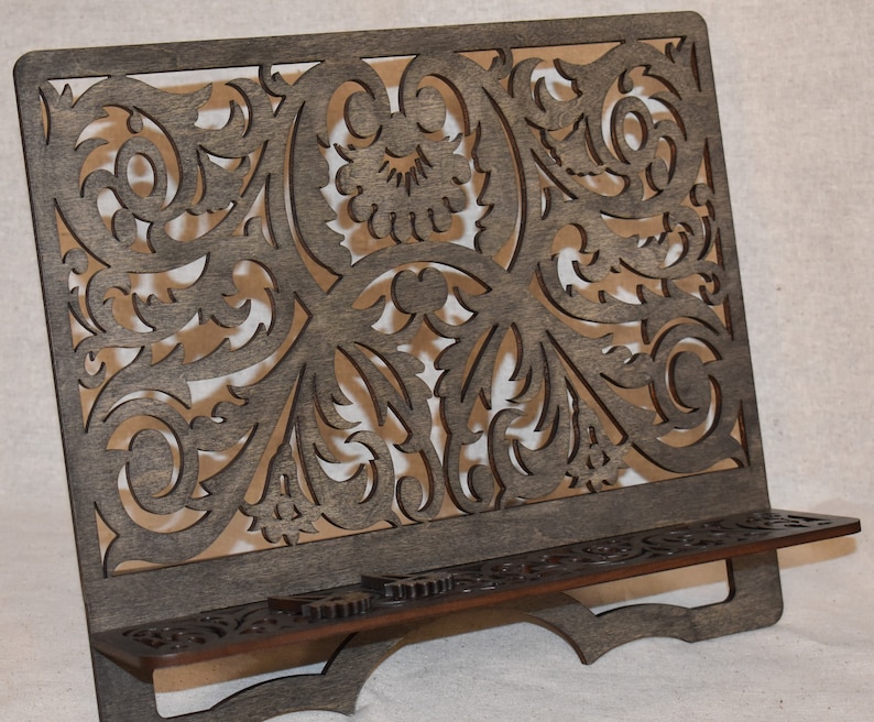 May include: A dark brown wooden book stand with a decorative, intricate, laser-cut design. The stand has a shelf for holding books or other items.