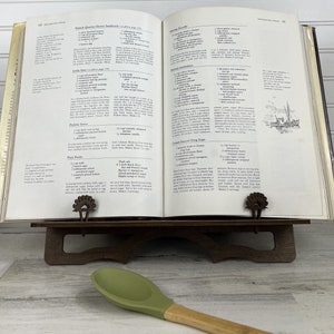 May include: Open cookbook with a brown wooden book stand and a green silicone spatula. The cookbook pages show recipes for French Quarter Oyster Sandwiches, Shrimp Jambalaya, and Cream Sauced Frog Legs.