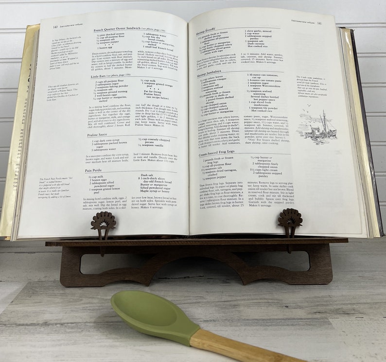 May include: Open cookbook with a brown wooden book stand and a green silicone spatula. The cookbook pages show recipes for French Quarter Oyster Sandwiches, Shrimp Jambalaya, and Cream Sauced Frog Legs.