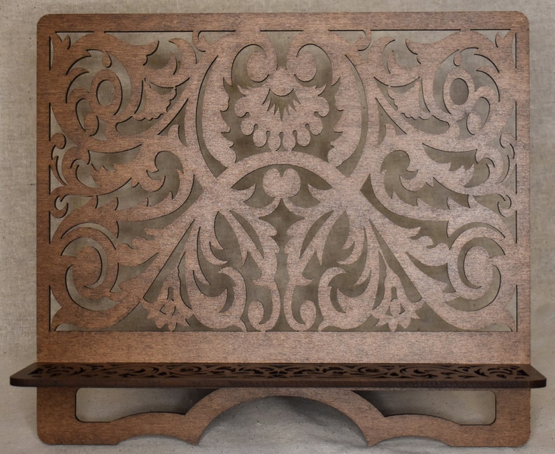 May include: A wooden music stand with a decorative, intricate, laser-cut design. The stand has a shelf and a curved base.