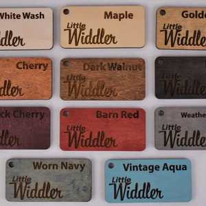 May include: Twelve wooden samples with the text "Little Widdler" in a cursive font. The samples are in various wood stains, including white wash, maple, golden oak, cherry, dark walnut, ebony, black cherry, barn red, weathered gray, worn navy, and vintage aqua.