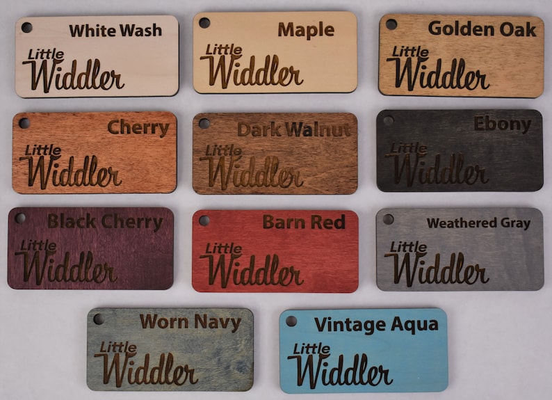 May include: Twelve wooden samples with the text "Little Widdler" in a cursive font. The samples are in various wood stains, including white wash, maple, golden oak, cherry, dark walnut, ebony, black cherry, barn red, weathered gray, worn navy, and vintage aqua.