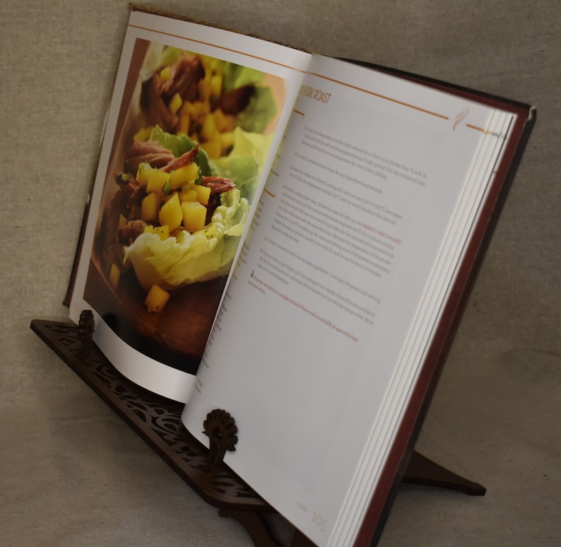 May include: A brown wooden cookbook stand with a decorative cut-out design. A cookbook is open on the stand, showing a recipe for "Pork Roast" with a photo of a lettuce wrap filled with pork and pineapple.