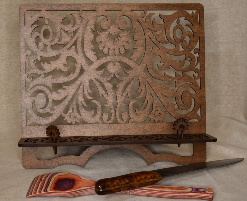 May include: A wooden cookbook stand with a decorative cutout design. The stand has a shelf and two metal page holders. The stand is brown and has a natural wood finish.