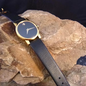 Op de afbeelding: Een goudkleurig polshorloge met een zwarte wijzerplaat en een enkele diamantmarkering op 12 uur. Het horloge heeft gouden wijzers en een zwarte leren band. Het horloge wordt getoond op een getextureerd rotsachtig oppervlak.