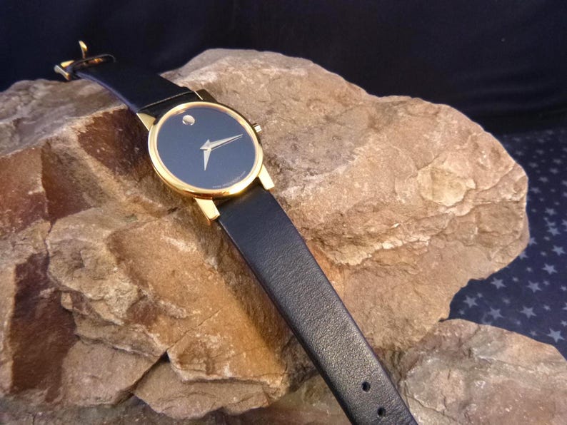 Op de afbeelding: Een goudkleurig polshorloge met een zwarte wijzerplaat en een enkele diamantmarkering op 12 uur. Het horloge heeft gouden wijzers en een zwarte leren band. Het horloge wordt getoond op een getextureerd rotsachtig oppervlak.