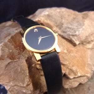 Op de afbeelding: Een goudkleurig polshorloge met een zwarte wijzerplaat en een zwarte leren band. Het horloge heeft een minimalistisch ontwerp met een enkele stip op 12 uur en gouden wijzers. Het horloge wordt gepresenteerd op een getextureerd rotsachtig oppervlak.