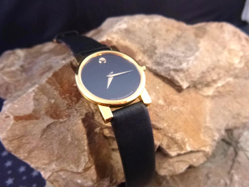 Op de afbeelding: Een goudkleurig polshorloge met een zwarte wijzerplaat en een zwarte leren band. Het horloge heeft een minimalistisch ontwerp met een enkele stip op 12 uur en gouden wijzers. Het horloge wordt gepresenteerd op een getextureerd rotsachtig oppervlak.