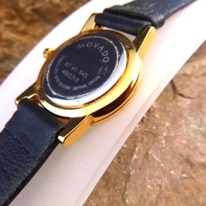 Op de afbeelding: Close-up van een Movado-horloge met een goudkleurige kast en een zwarte leren band. De achterkant van het horloge is zichtbaar, met het Movado-logo en modelinformatie. Het horloge is waterbestendig met saffierkristal.