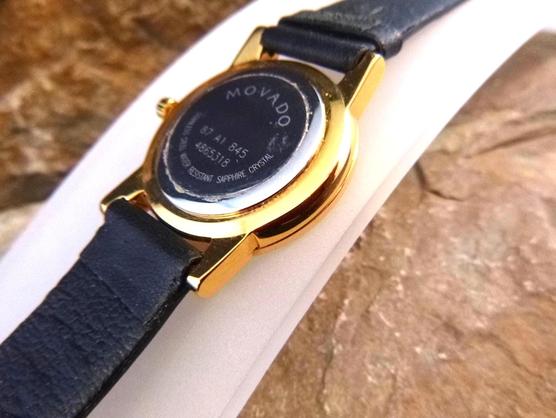 Op de afbeelding: Close-up van een Movado-horloge met een goudkleurige kast en een zwarte leren band. De achterkant van het horloge is zichtbaar, met het Movado-logo en modelinformatie. Het horloge is waterbestendig met saffierkristal.