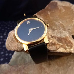Op de afbeelding: Een goudkleurig polshorloge met een zwarte leren band en een donkerblauwe wijzerplaat. Het horloge heeft een minimalistisch ontwerp met &eacute;&eacute;n zilverkleurige wijzer. Het horloge ligt op een getextureerd rotsachtig oppervlak.