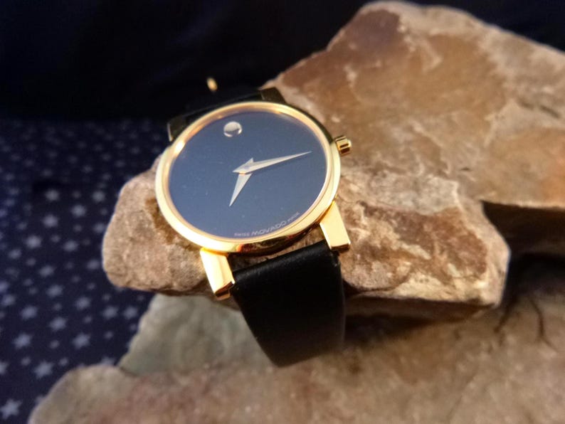 Op de afbeelding: Een goudkleurig polshorloge met een zwarte leren band en een donkerblauwe wijzerplaat. Het horloge heeft een minimalistisch ontwerp met &eacute;&eacute;n zilverkleurige wijzer. Het horloge ligt op een getextureerd rotsachtig oppervlak.