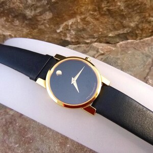 Op de afbeelding: Een Movado horloge met een zwarte wijzerplaat en een goudkleurige kast. Het horloge heeft een zwarte leren band en een minimalistisch ontwerp met een enkele stip op 12 uur. Het horloge wordt gepresenteerd op een witte ondergrond.