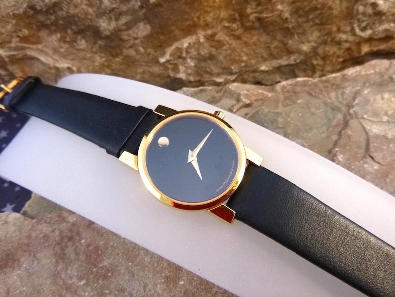 Op de afbeelding: Een Movado horloge met een zwarte wijzerplaat en een goudkleurige kast. Het horloge heeft een zwarte leren band en een minimalistisch ontwerp met een enkele stip op 12 uur. Het horloge wordt gepresenteerd op een witte ondergrond.