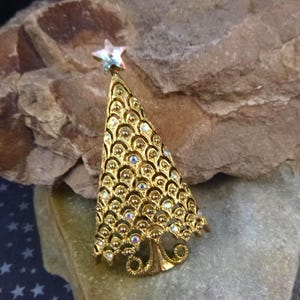 May include: Gold-toned Christmas tree brooch with a star on top. The tree is adorned with small, iridescent rhinestones and a textured pattern. A festive accessory for the holiday season.