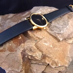 Op de afbeelding: Een zwart wijzerplaat horloge met een goudkleurige kast en een zwarte leren band. Het horloge heeft een minimalistisch ontwerp met een enkele stip op 12 uur. Het horloge wordt getoond op een getextureerde, lichtbruine rots.