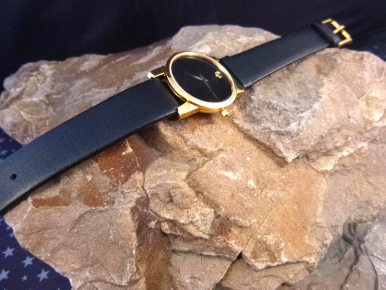 Op de afbeelding: Een zwart wijzerplaat horloge met een goudkleurige kast en een zwarte leren band. Het horloge heeft een minimalistisch ontwerp met een enkele stip op 12 uur. Het horloge wordt getoond op een getextureerde, lichtbruine rots.