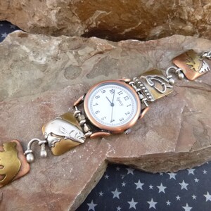 May include: A silver and copper watch with a white face and black Roman numeral hour markers. The watch strap is made of metal and features a series of decorative metal charms with images of fish, trees, and other nature-inspired designs.