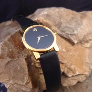 Op de afbeelding: Een polshorloge met een zwarte wijzerplaat, een goudkleurige kast en een zwarte leren band. Het horloge heeft een minimalistisch ontwerp met een enkele stip op 12 uur en gouden wijzers. Het horloge ligt op een getextureerd rotsachtig oppervlak.