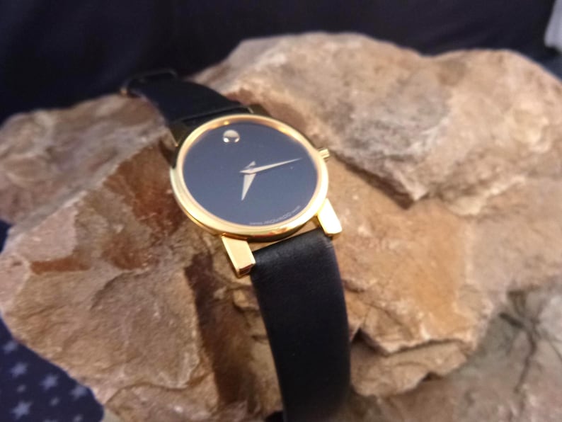 Op de afbeelding: Een polshorloge met een zwarte wijzerplaat, een goudkleurige kast en een zwarte leren band. Het horloge heeft een minimalistisch ontwerp met een enkele stip op 12 uur en gouden wijzers. Het horloge ligt op een getextureerd rotsachtig oppervlak.