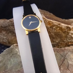 Op de afbeelding: Een horloge met een zwarte leren band, een goudkleurige kast en een donkerblauwe wijzerplaat. Het horloge heeft een enkele stip op 12 uur en een goudkleurige wijzer. Het horloge wordt gepresenteerd op een wit oppervlak.