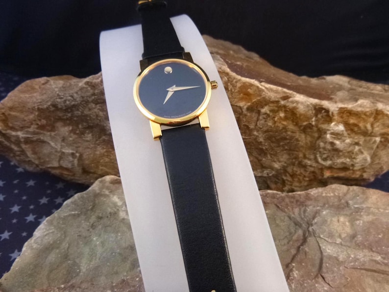 Op de afbeelding: Een horloge met een zwarte leren band, een goudkleurige kast en een donkerblauwe wijzerplaat. Het horloge heeft een enkele stip op 12 uur en een goudkleurige wijzer. Het horloge wordt gepresenteerd op een wit oppervlak.
