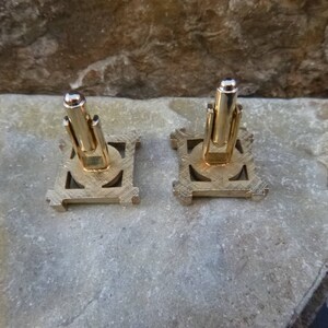 May include: A pair of gold-toned cufflinks with a geometric design. The cufflinks have a square shape with a circular center.