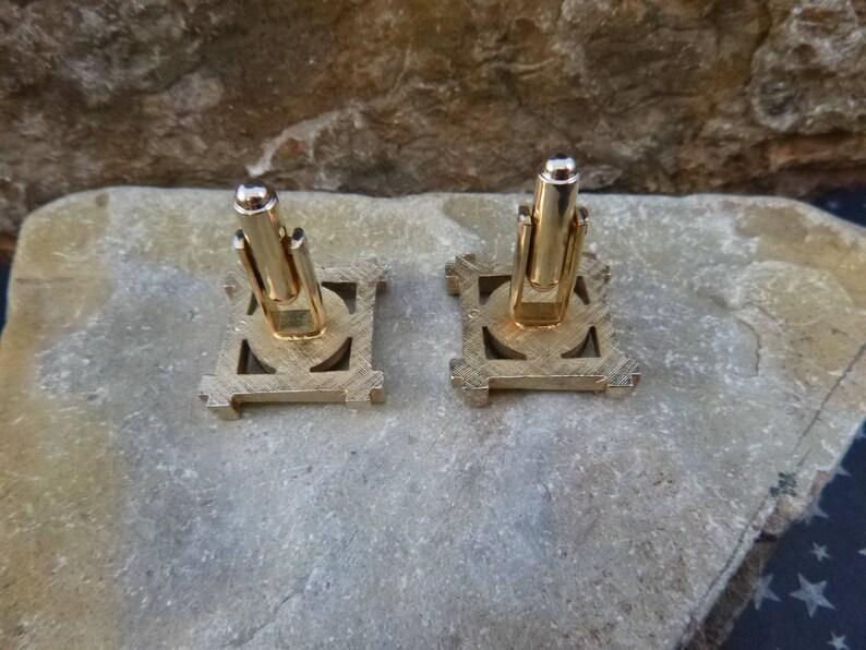 May include: A pair of gold-toned cufflinks with a geometric design. The cufflinks have a square shape with a circular center.
