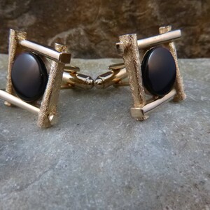 May include: A pair of gold cufflinks with black oval stones set in a square frame design.