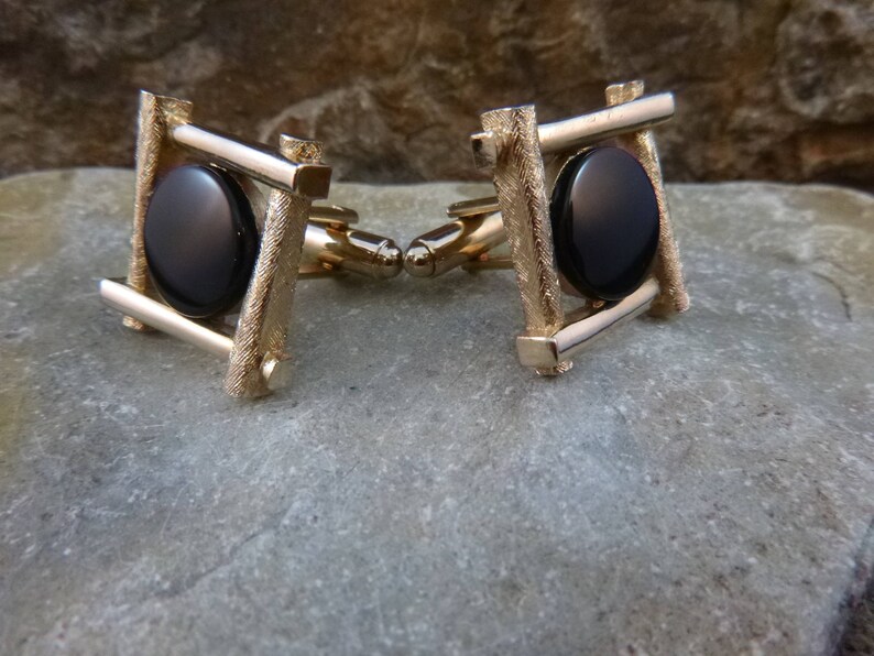 May include: A pair of gold cufflinks with black oval stones set in a square frame design.