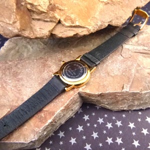 Op de afbeelding: Een goudkleurig Movado-horloge met een zwarte leren band. De wijzerplaat van het horloge is niet zichtbaar, maar de achterkant van het horloge is zichtbaar, met het Movado-logo en andere tekst. Het horloge ligt op een rotsachtig oppervlak.