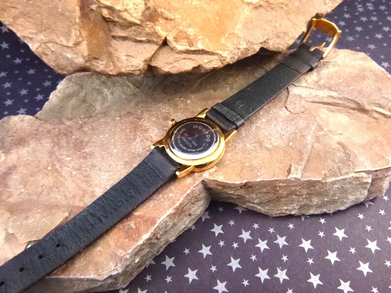 Op de afbeelding: Een goudkleurig Movado-horloge met een zwarte leren band. De wijzerplaat van het horloge is niet zichtbaar, maar de achterkant van het horloge is zichtbaar, met het Movado-logo en andere tekst. Het horloge ligt op een rotsachtig oppervlak.