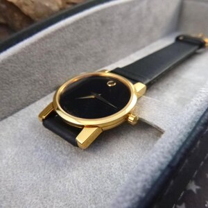 Op de afbeelding: Een goudkleurig polshorloge met een zwarte wijzerplaat en een zwarte leren band. Het horloge is geplaatst in een grijze, gewatteerde etui. Het horloge heeft een minimalistisch ontwerp met een enkele goudkleurige stip op 12 uur.