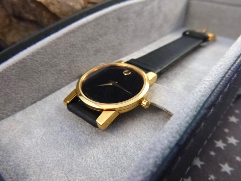 Op de afbeelding: Een goudkleurig polshorloge met een zwarte wijzerplaat en een zwarte leren band. Het horloge is geplaatst in een grijze, gewatteerde etui. Het horloge heeft een minimalistisch ontwerp met een enkele goudkleurige stip op 12 uur.