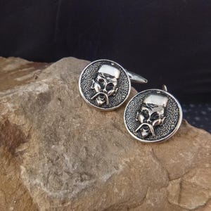 May include: A pair of silver cufflinks with a detailed design of a man's face with a mustache and a hat. The cufflinks are on a gray rock.