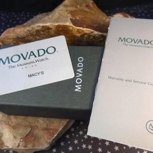 Op de afbeelding: Een stilleven met een Movado horlogedoos, garantiekaart en een Macy's kaart. De doos is donkergrijs met het Movado logo. De garantiekaart is lichtgrijs met het Movado logo en tekst. De Macy's kaart is wit met het Movado logo en de Macy's tekst.