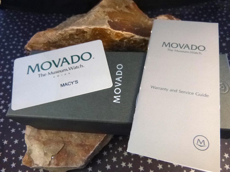 Op de afbeelding: Een stilleven met een Movado horlogedoos, garantiekaart en een Macy's kaart. De doos is donkergrijs met het Movado logo. De garantiekaart is lichtgrijs met het Movado logo en tekst. De Macy's kaart is wit met het Movado logo en de Macy's tekst.
