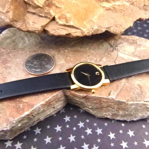 Op de afbeelding: Een goudkleurig polshorloge met een zwarte wijzerplaat en een zwarte leren band. Het horloge wordt getoond op een rotsachtig oppervlak naast een munt. Het horloge heeft een minimalistisch ontwerp.