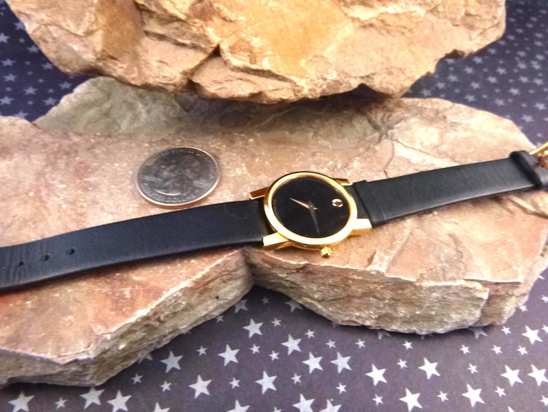 Op de afbeelding: Een goudkleurig polshorloge met een zwarte wijzerplaat en een zwarte leren band. Het horloge wordt getoond op een rotsachtig oppervlak naast een munt. Het horloge heeft een minimalistisch ontwerp.
