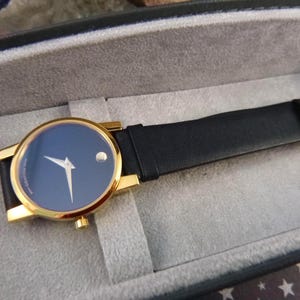 Op de afbeelding: Goudkleurig horloge met een marineblauwe wijzerplaat en een zwarte leren band. Het horloge heeft een minimalistisch ontwerp met gouden wijzers en een enkele stip op 12 uur. Het horloge wordt gepresenteerd in een grijze doos.