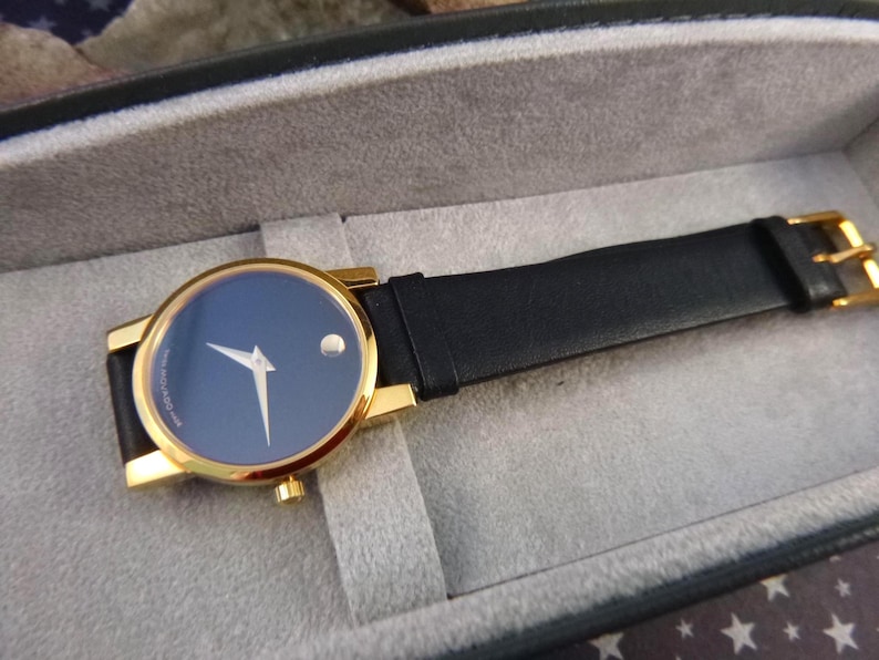 Op de afbeelding: Goudkleurig horloge met een marineblauwe wijzerplaat en een zwarte leren band. Het horloge heeft een minimalistisch ontwerp met gouden wijzers en een enkele stip op 12 uur. Het horloge wordt gepresenteerd in een grijze doos.