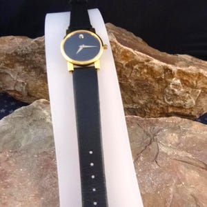 Op de afbeelding: Een zwart leren horloge met een goudkleurige bezel en een donkerblauwe wijzerplaat. Het horloge heeft een kleine diamant op 12 uur en een zwarte leren band. Het horloge wordt getoond op een witte standaard tegen een natuurlijke rotsachtergrond.