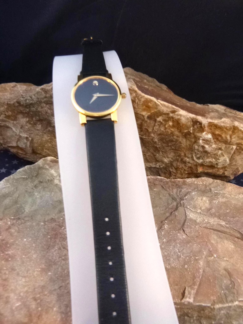 Op de afbeelding: Een zwart leren horloge met een goudkleurige bezel en een donkerblauwe wijzerplaat. Het horloge heeft een kleine diamant op 12 uur en een zwarte leren band. Het horloge wordt getoond op een witte standaard tegen een natuurlijke rotsachtergrond.